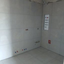 Snowbud Bartosz Śnieżewski - Wykończona łazienka z jasnymi płytkami, widoczne punkty instalacji wodnej i elektrycznej, oraz skrzynka bezpiecznikowa. Surowy strop z instalacjami.