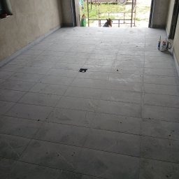 Snowbud Bartosz Śnieżewski - Wykończona podłoga z szarych płytek w pomieszczeniu z surowymi ścianami. Na zewnątrz widoczny samochód i pies.