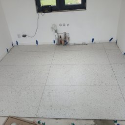 Snowbud Bartosz Śnieżewski - Ułożone płytki podłogowe w łazience, zabezpieczone niebieską taśmą. Widoczne przyłącza wodne i okno. Płytki w kolorze białym z drobnymi, kolorowymi elementami.