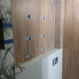 Snowbud Bartosz Śnieżewski - Montaż drewnopodobnych paneli ściennych w łazience, z użyciem klipsów poziomujących. Widoczne płytki z motywem roślinnym i zabudowa stelaża WC.