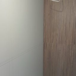 Snowbud Bartosz Śnieżewski - Ściana wykończona w połowie: gładka, biała powierzchnia z lewej, dekoracyjne panele drewniane z prawej. Widoczne przewody elektryczne u góry.