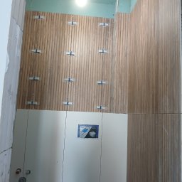 Snowbud Bartosz Śnieżewski - Montaż drewnopodobnych płytek ściennych w łazience z użyciem krzyżyków dystansowych. Widoczne białe szafki i instalacja podtynkowa WC. Etap prac wykończeniowych.