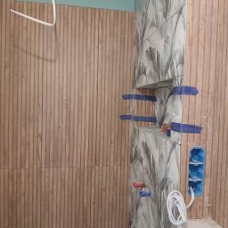 Snowbud Bartosz Śnieżewski - Ściana z płytek imitujących drewno i płytek z motywem liści, z wnęką i puszką elektryczną. Widoczne niebieskie taśmy zabezpieczające.