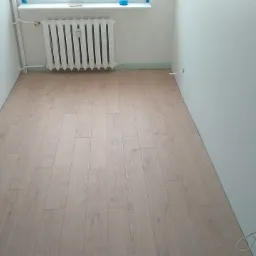 Jasne panele podłogowe ułożone w pustym pokoju z białymi ścianami i grzejnikiem pod oknem, widoczne kable elektryczne wystające ze ściany.