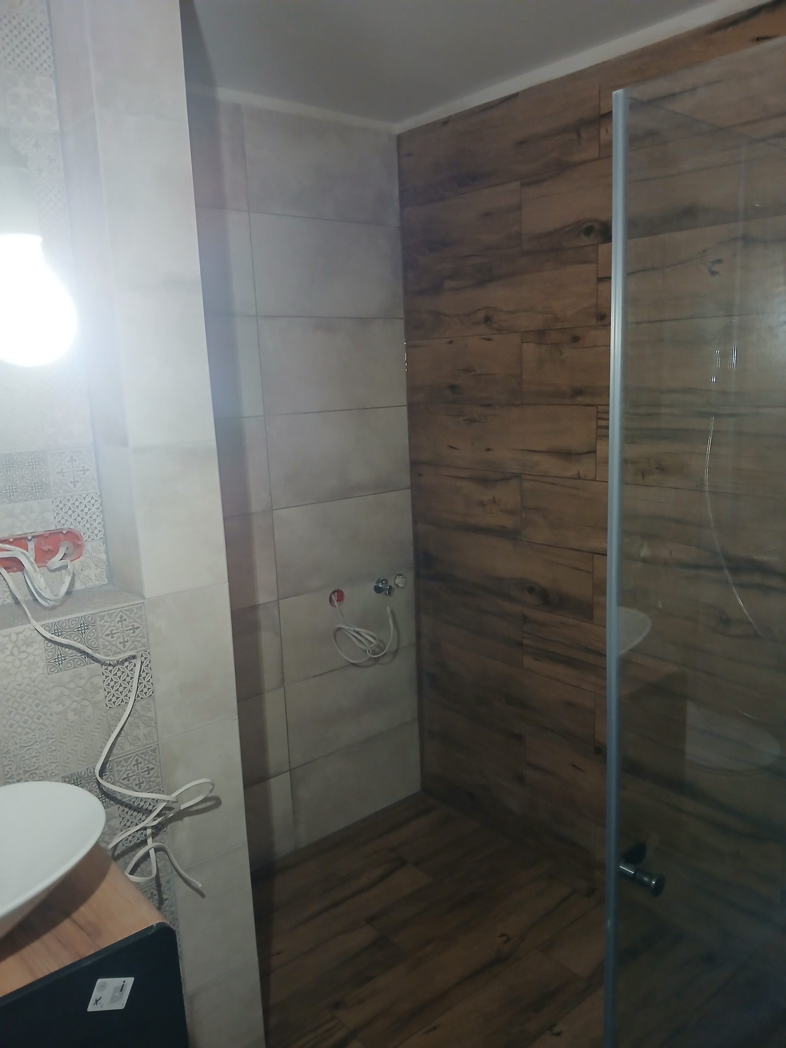 Wykończona łazienka z kabiną prysznicową i płytkami imitującymi drewno. Widoczne przyłącza wodne i umywalka z szafką. Oświetlenie górne.