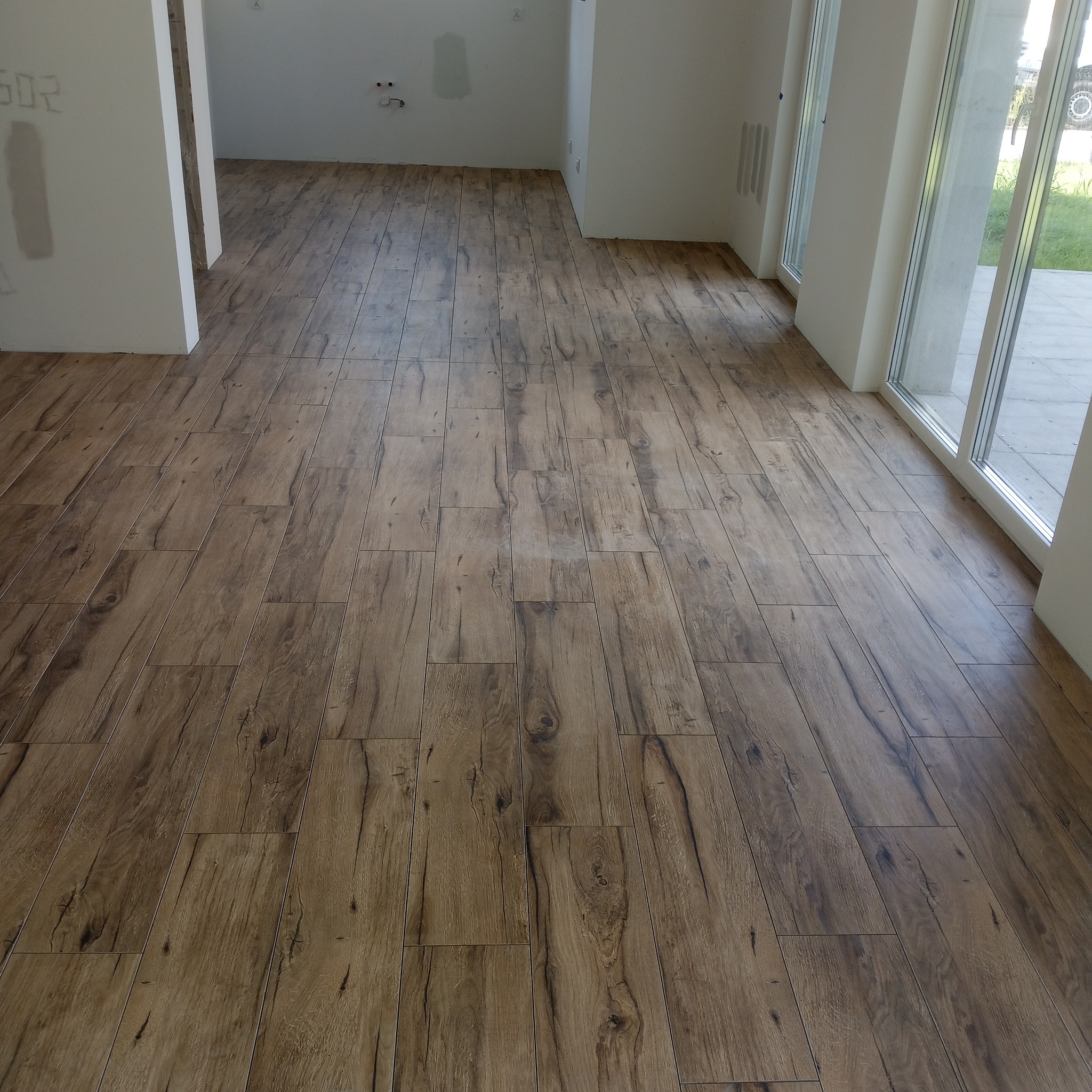 Ukończona podłoga z płytek imitujących drewno w jasnym, przestronnym wnętrzu. Widoczne duże okna i surowe ściany w trakcie wykańczania.