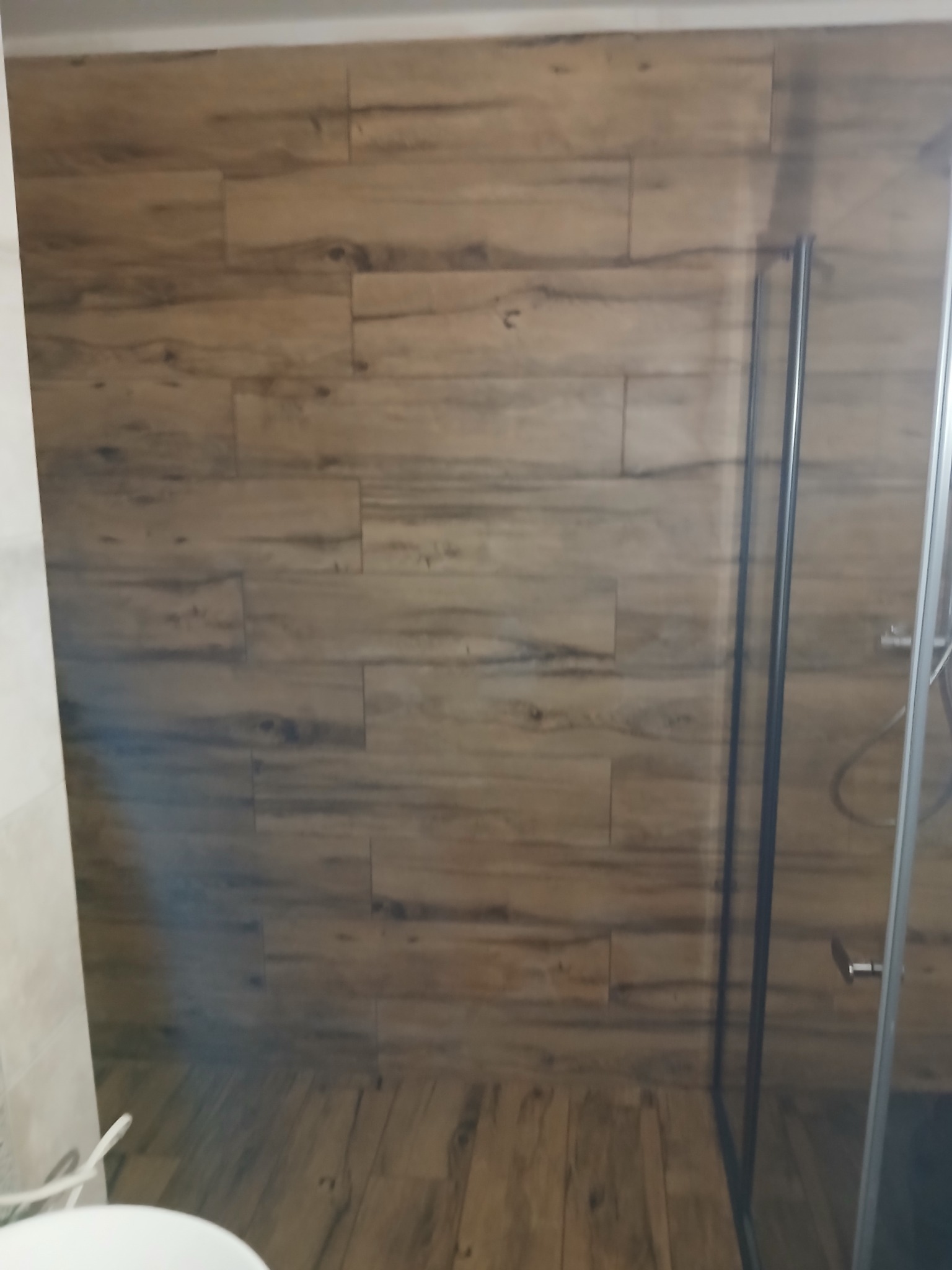 Kabina prysznicowa z drewnopodobnymi płytkami na ścianach i podłodze, widok z boku, minimalistyczny design, szklane drzwi z czarnymi profilami.