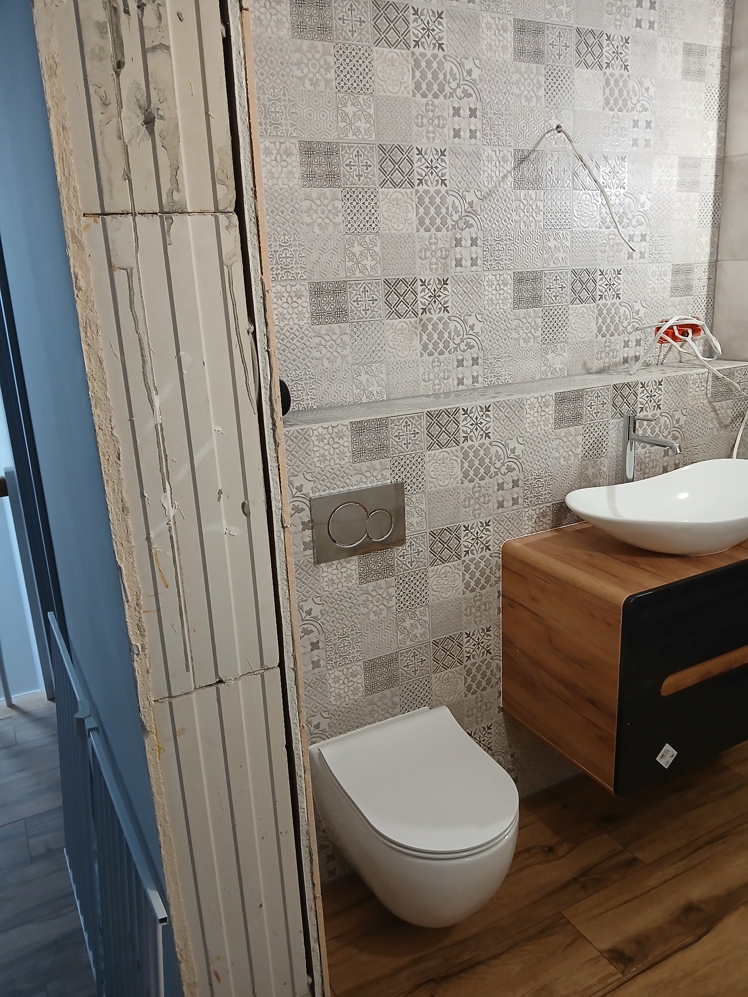 Nowoczesna łazienka w trakcie wykańczania: biała umywalka nablatowa, wisząca toaleta i dekoracyjne płytki ścienne w odcieniach szarości. Widoczna ściana z pustaków.