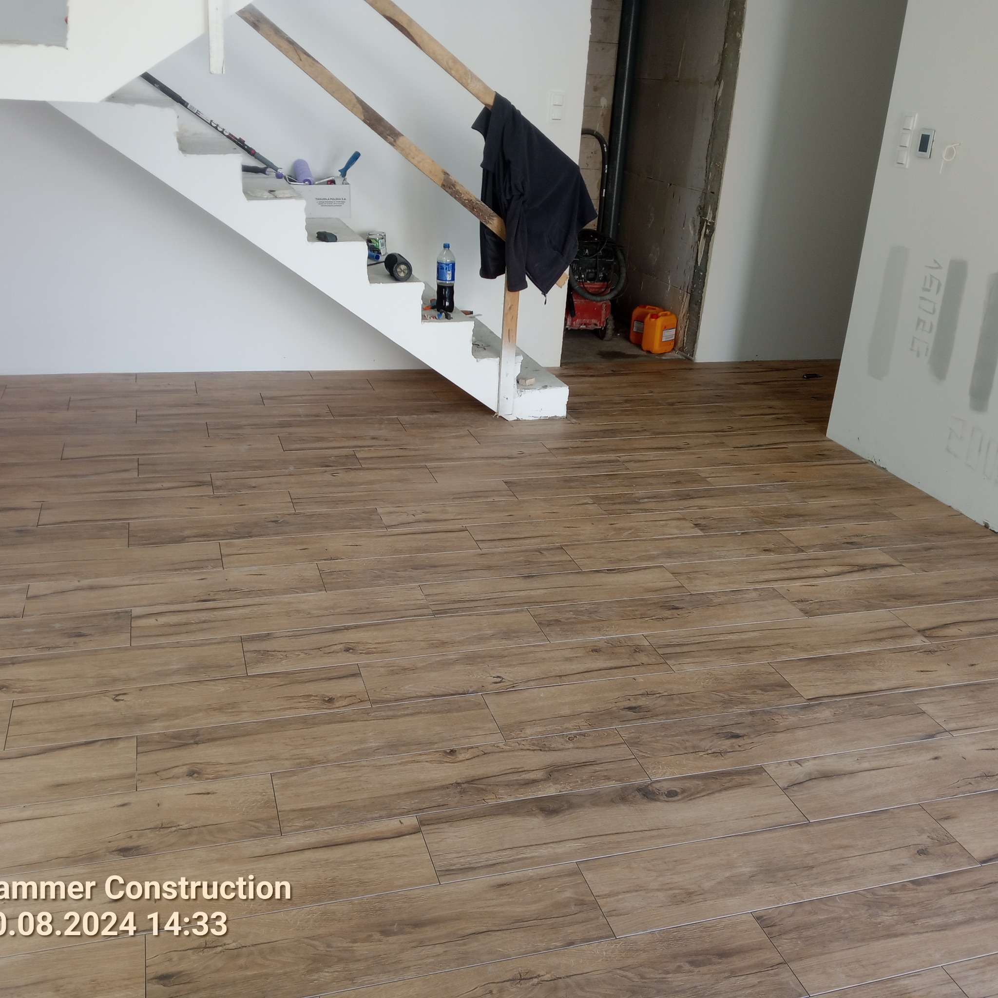 Ukończona podłoga z płytek imitujących drewno, ułożona w nowym domu. Widoczne schody w trakcie wykańczania i narzędzia malarskie.