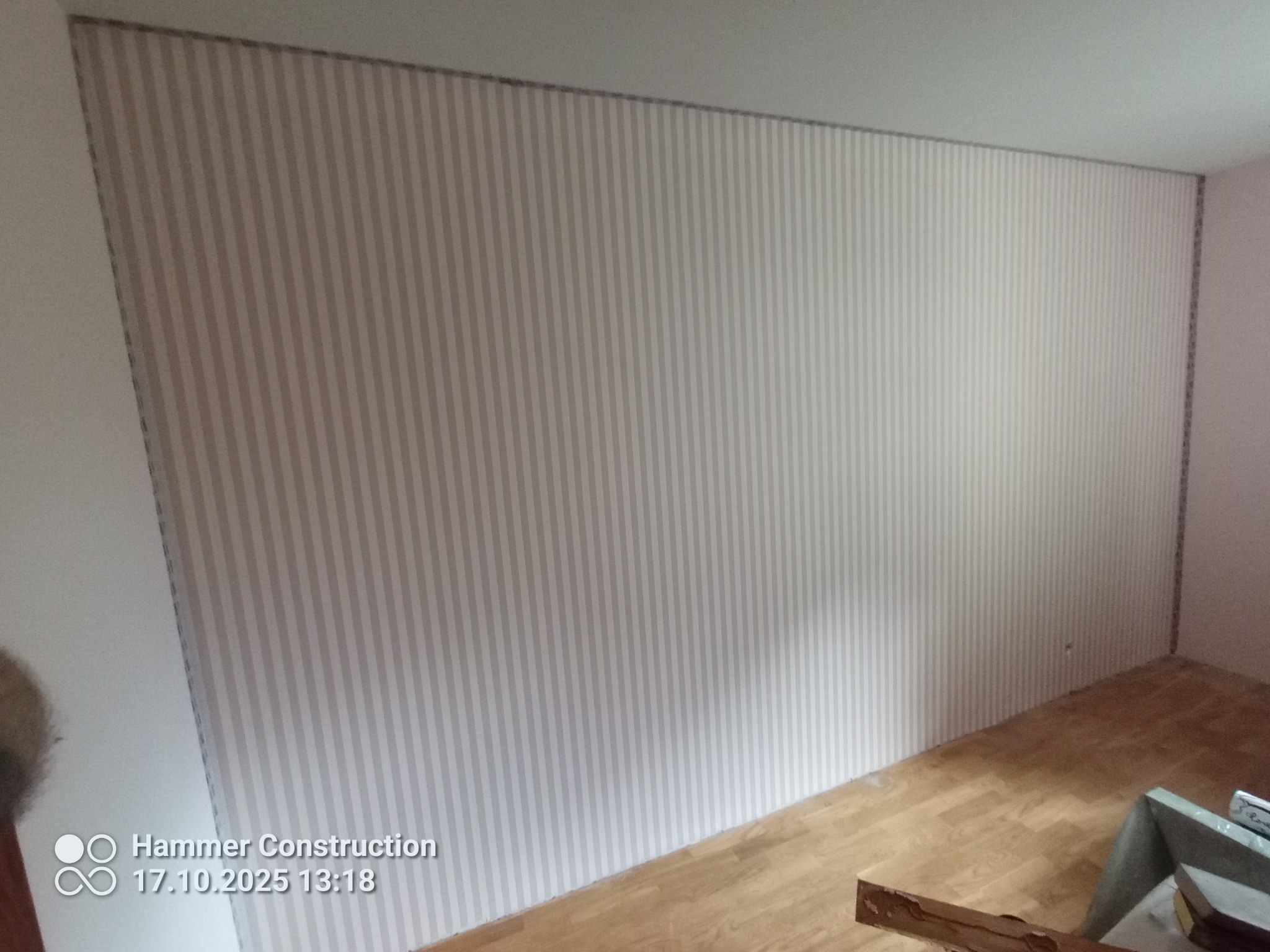 Ściana po tapetowaniu w pionowe pasy, widoczna podłoga z paneli. Fragment drabiny w prawym dolnym rogu. Jasne, naturalne oświetlenie.