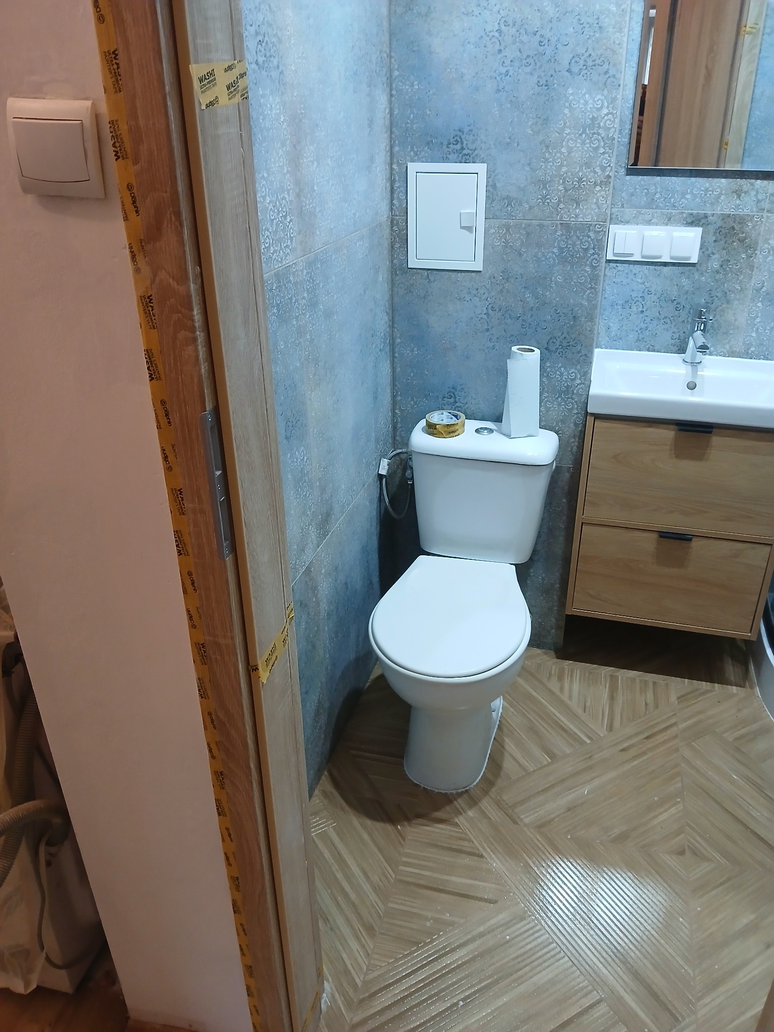 Wykończona łazienka z toaletą, umywalką z szafką i szaro-niebieskimi płytkami. Na podłodze brązowe płytki imitujące drewno. Drzwi zabezpieczone taśmą malarską.