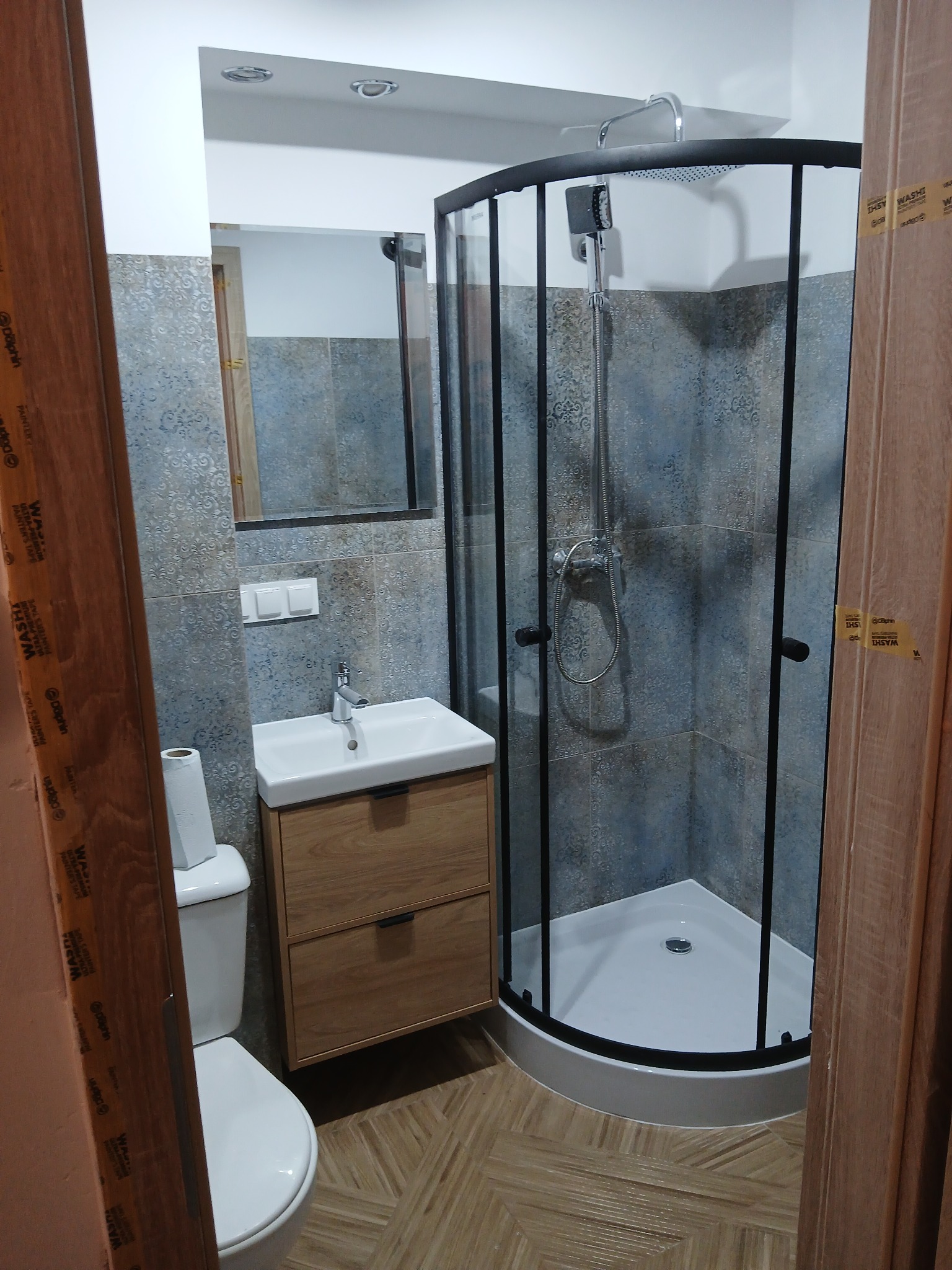 Nowoczesna łazienka po remoncie: kabina prysznicowa z czarnymi profilami, umywalka z szafką w odcieniach drewna, toaleta i płytki w odcieniach szarości i błękitu.