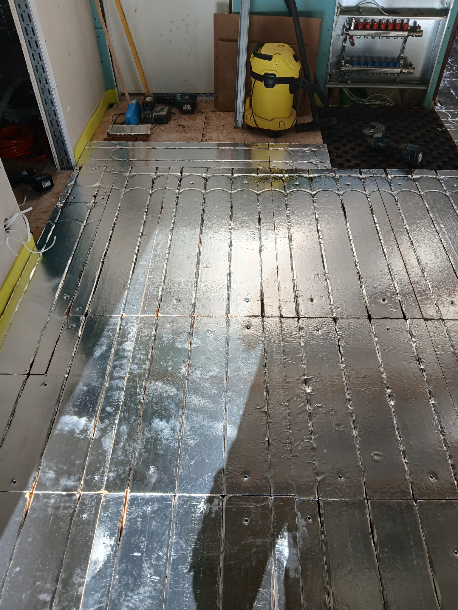 Rozłożona folia aluminiowa na podłodze, przygotowana pod ogrzewanie podłogowe. Widoczne narzędzia i elementy instalacji. Żółty odkurzacz przemysłowy w tle.