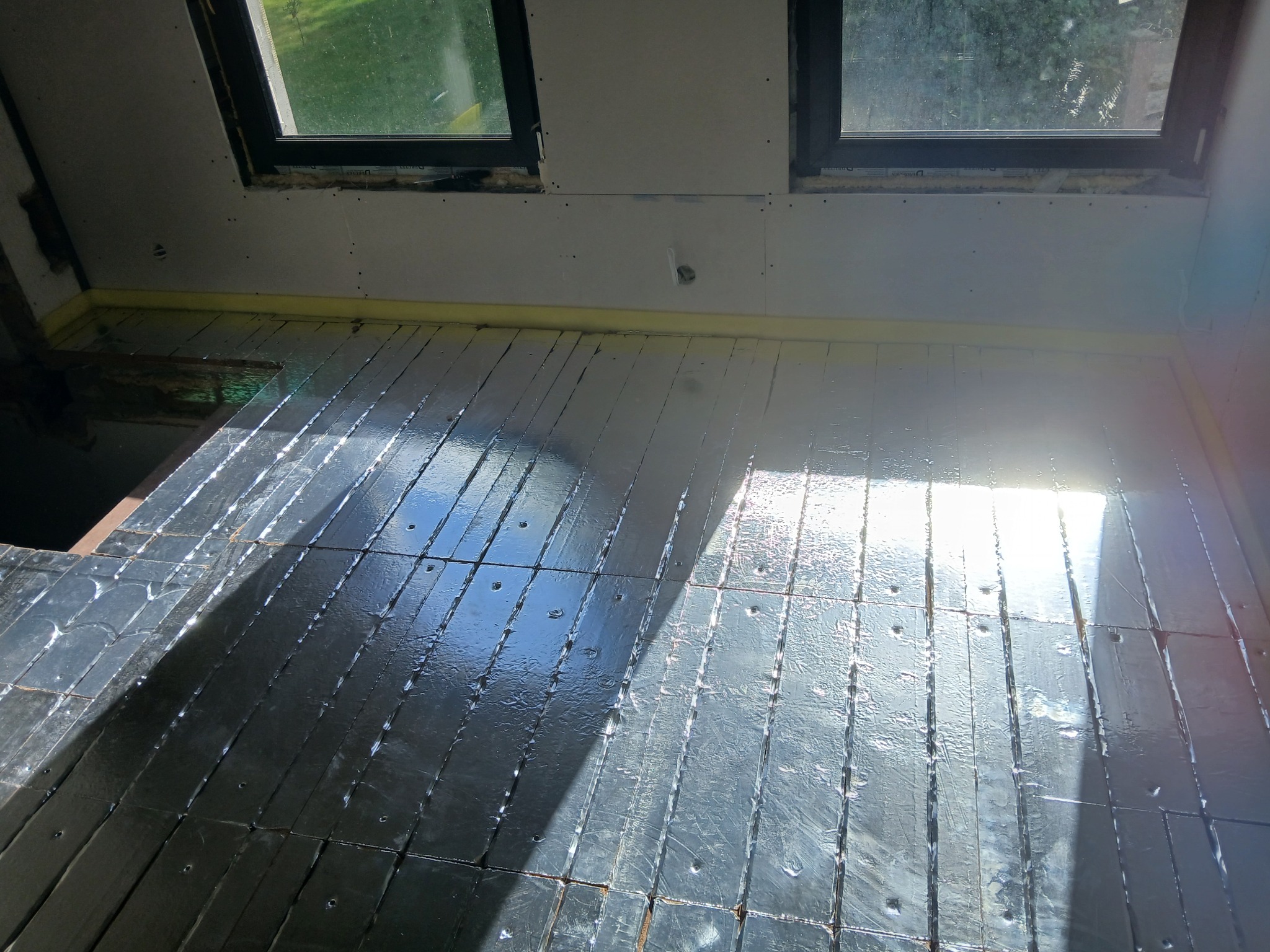 Podłoga na poddaszu z izolacją termiczną, przygotowana pod wylewkę. Widoczne okna dachowe i płyty kartonowo-gipsowe na ścianach. Jasne światło słoneczne.