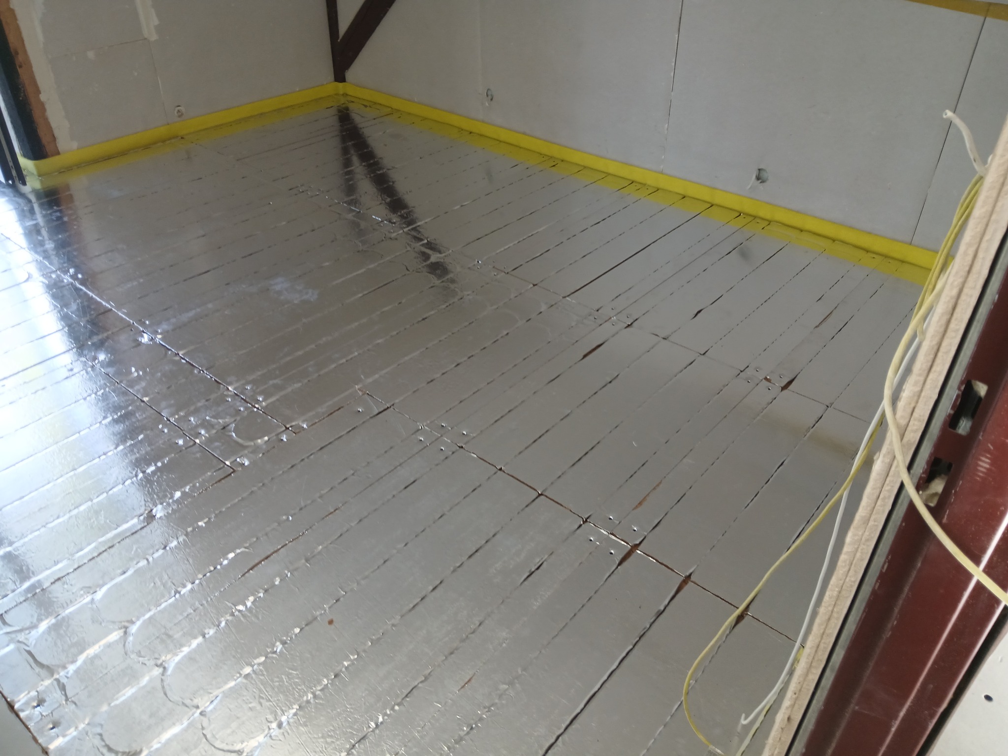 Rozłożona folia aluminiowa z widocznymi liniami ogrzewania podłogowego, zabezpieczona żółtą taśmą przy ścianach w pomieszczeniu w trakcie prac wykończeniowych.