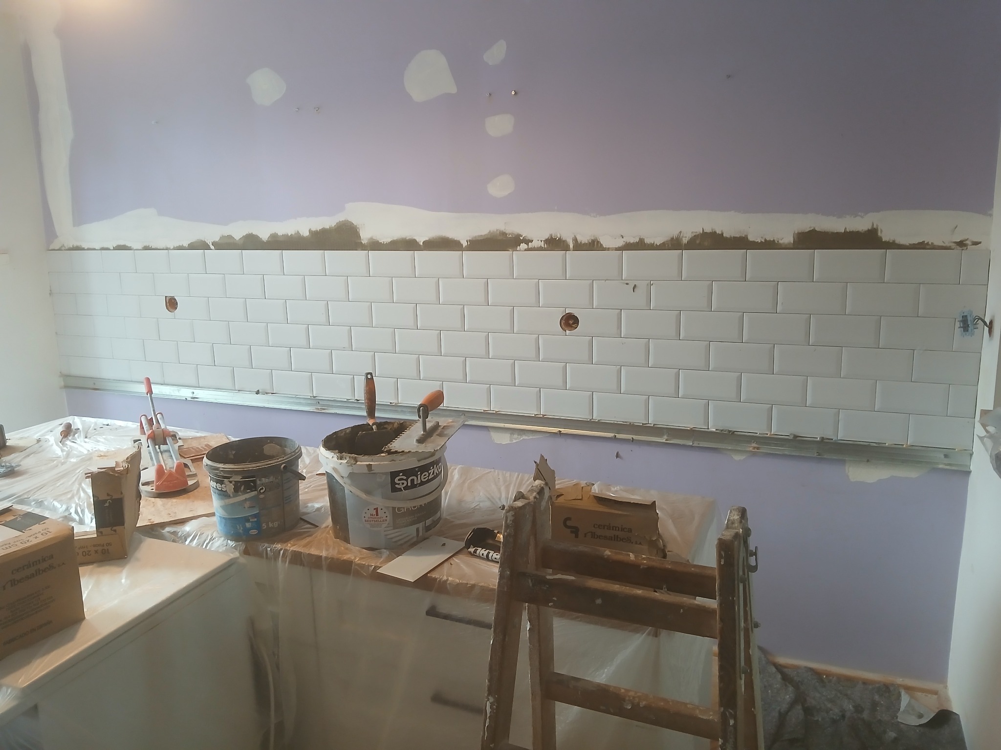 Białe, prostokątne płytki ułożone na ścianie w trakcie prac. Widoczne narzędzia, wiadro z klejem i drabina. Ściana pomalowana na fioletowo, z widocznymi śladami po gładzi.