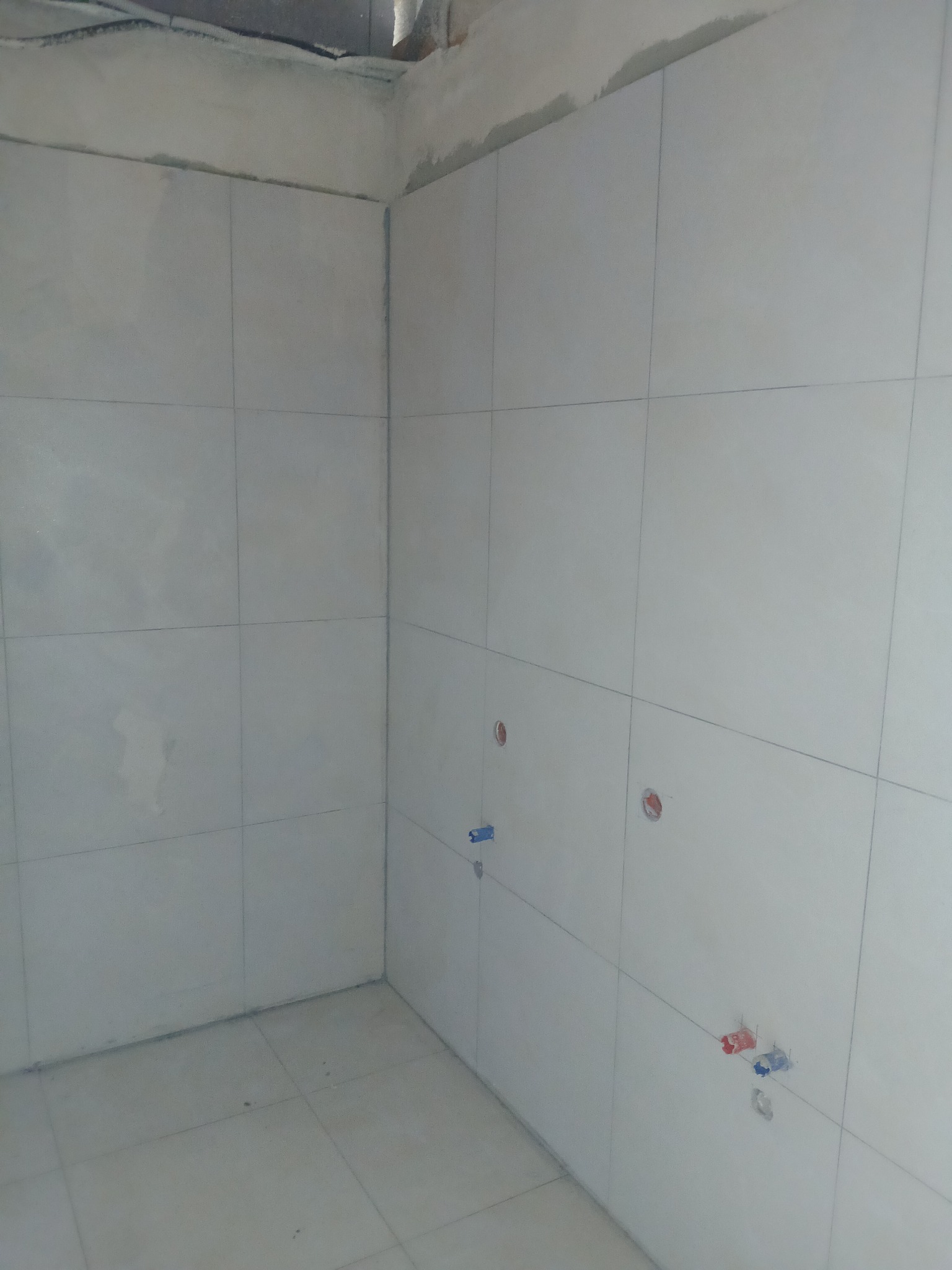 Wykończona, jasna łazienka z białymi, kwadratowymi płytkami na ścianach i podłodze. Widoczne otwory na instalacje wodne. Surowy stan wykończenia sufitu.