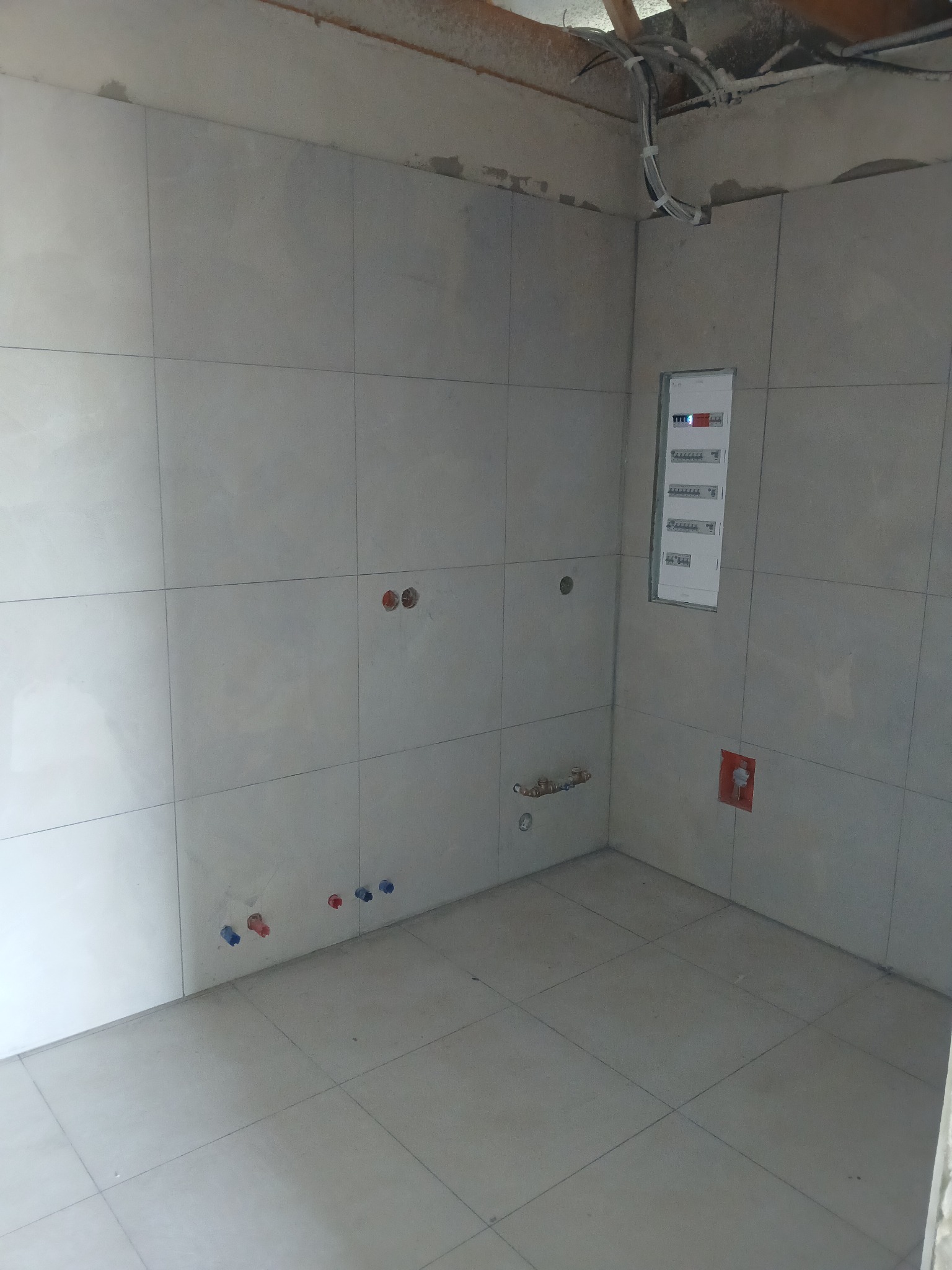 Wykończona łazienka z jasnymi płytkami, widoczne punkty instalacji wodnej i elektrycznej, oraz skrzynka bezpiecznikowa. Surowy strop z instalacjami.