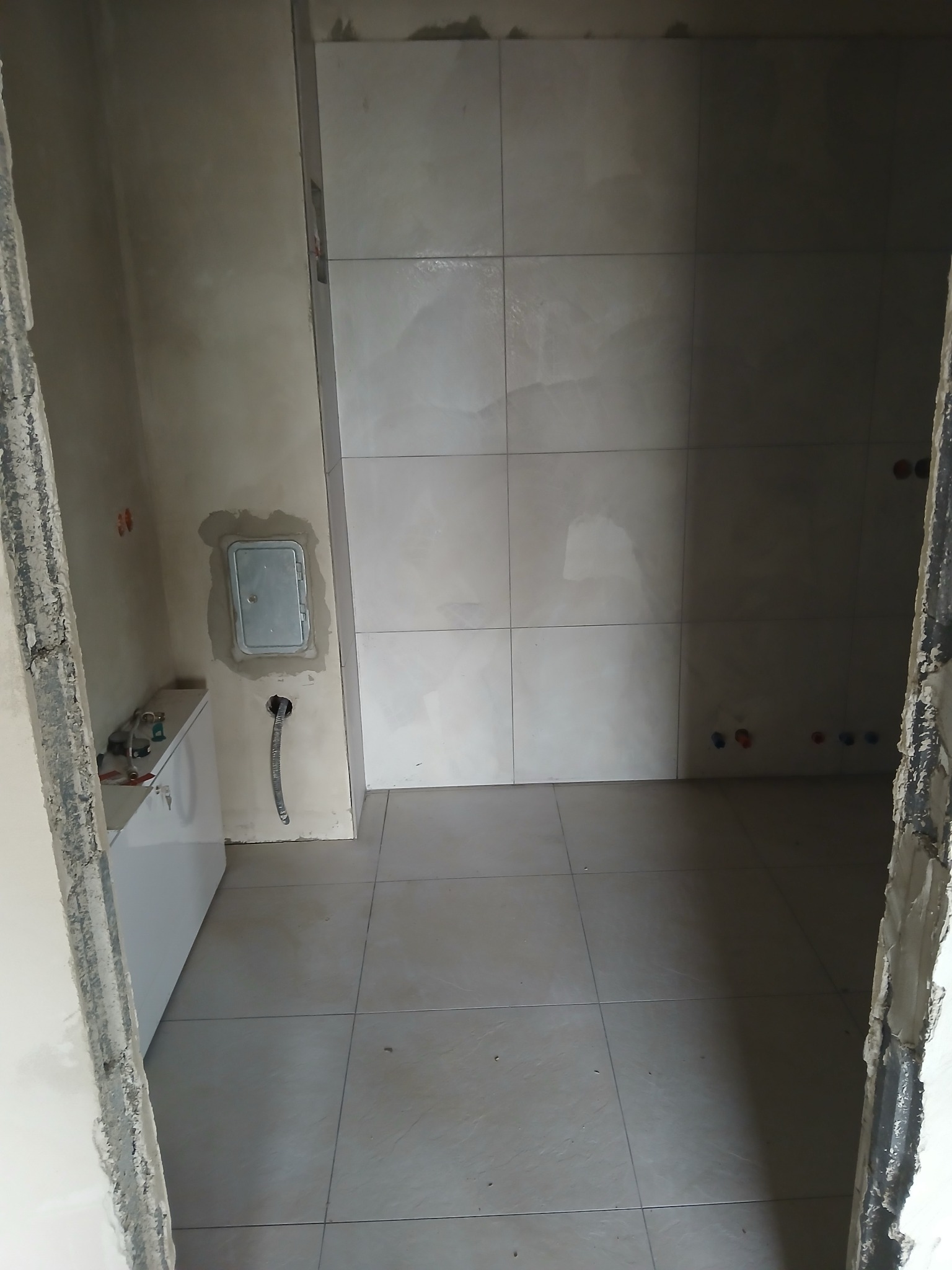 Wnętrze łazienki w trakcie prac: ułożone płytki na ścianach i podłodze, widoczna instalacja wodna, surowy stan ścian przy wejściu.