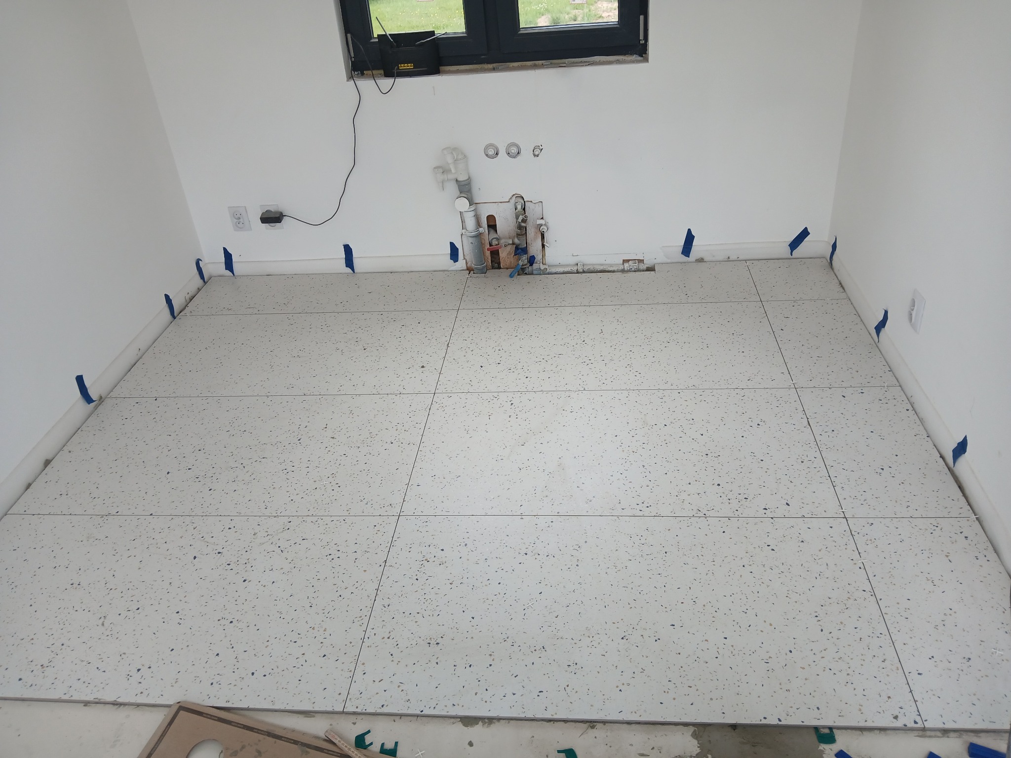 Ułożone płytki podłogowe w łazience, zabezpieczone niebieską taśmą. Widoczne przyłącza wodne i okno. Płytki w kolorze białym z drobnymi, kolorowymi elementami.