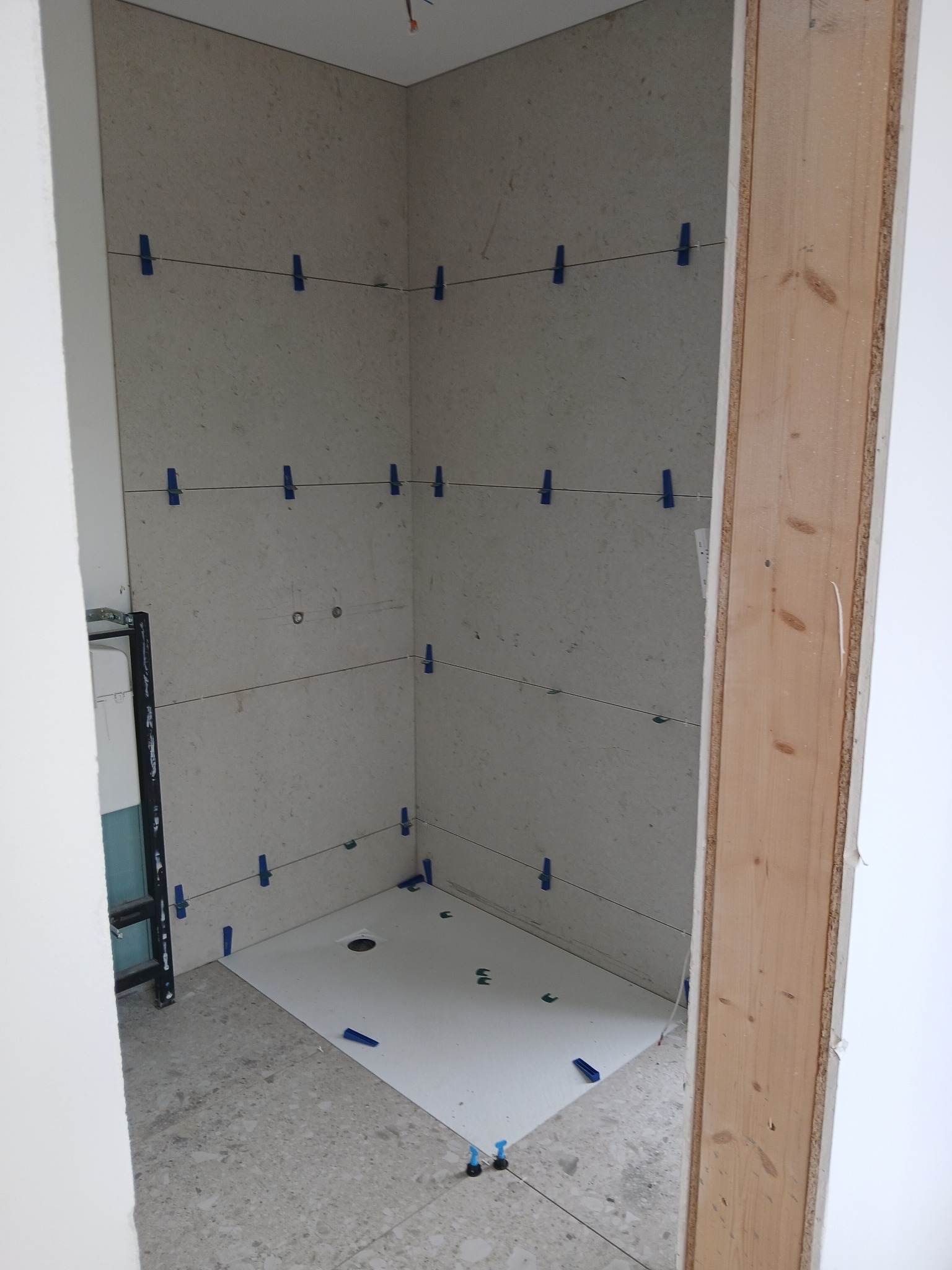 Wnętrze łazienki w trakcie prac. Widoczne płytki na ścianach, zabezpieczone niebieskimi klipsami, oraz biała podłoga z odpływem. Fragment drewnianej futryny w kadrze.