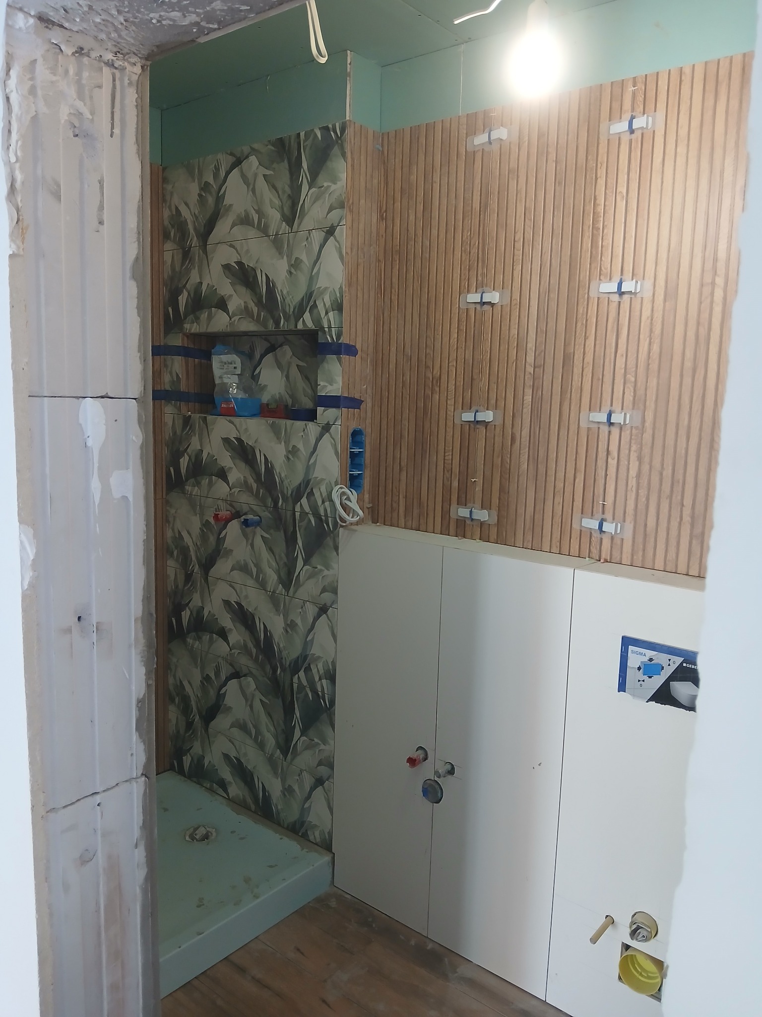 Łazienka w trakcie prac: montaż płytek z motywem roślinnym i drewnopodobnych paneli. Widoczne elementy instalacji i stelaż podtynkowy WC. Oświetlenie sufitowe.