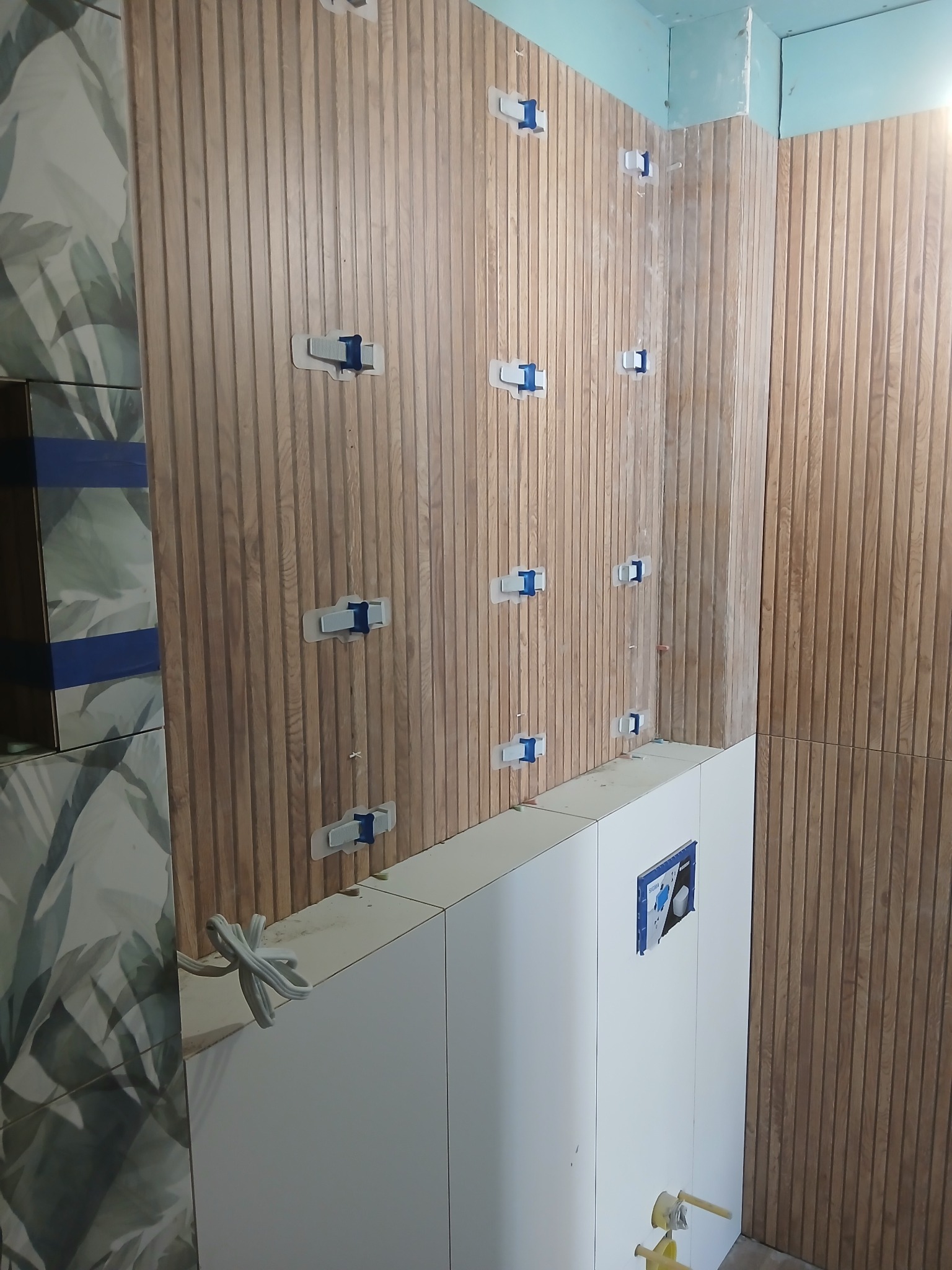 Montaż drewnopodobnych paneli ściennych w łazience, z użyciem klipsów poziomujących. Widoczne płytki z motywem roślinnym i zabudowa stelaża WC.