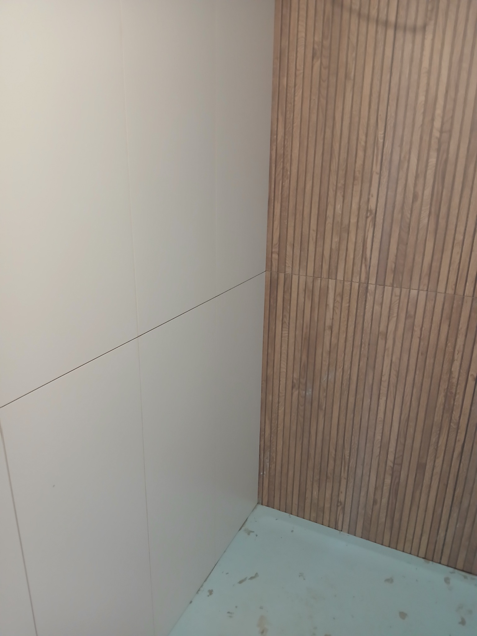Wykończona ściana z płytek imitujących drewno, sąsiadująca z gładką, jasną ścianą. Widoczny fragment podłogi. Minimalistyczny design.