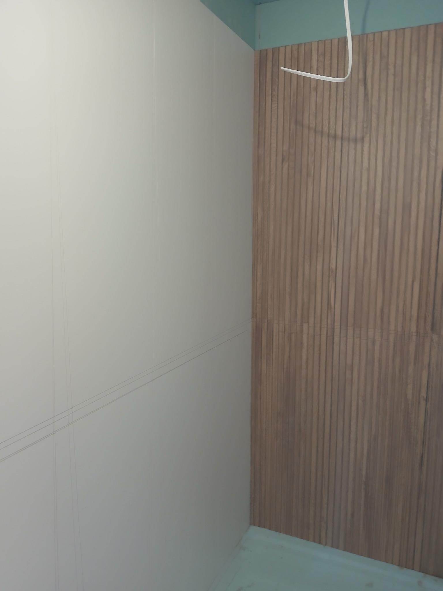 Ściana wykończona w połowie: gładka, biała powierzchnia z lewej, dekoracyjne panele drewniane z prawej. Widoczne przewody elektryczne u góry.