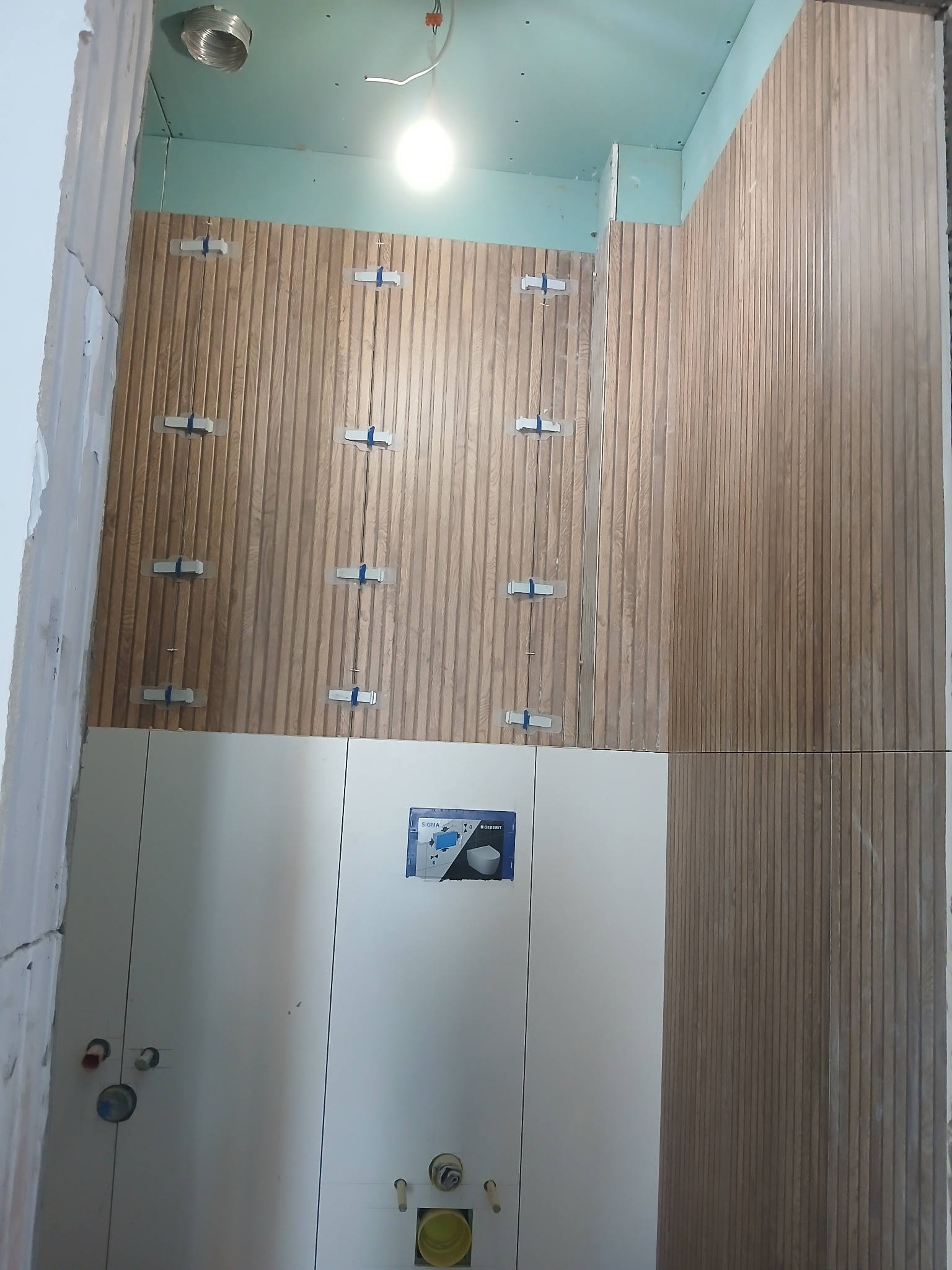 Montaż drewnopodobnych płytek ściennych w łazience z użyciem krzyżyków dystansowych. Widoczne białe szafki i instalacja podtynkowa WC. Etap prac wykończeniowych.