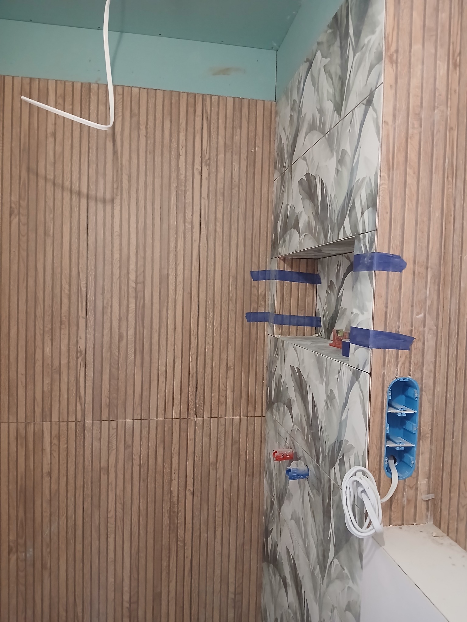 Ściana z płytek imitujących drewno i płytek z motywem liści, z wnęką i puszką elektryczną. Widoczne niebieskie taśmy zabezpieczające.