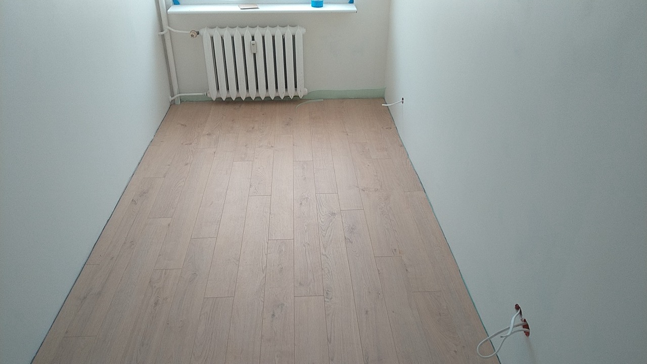 Jasne panele podłogowe ułożone w pustym pokoju z białymi ścianami i grzejnikiem pod oknem, widoczne kable elektryczne wystające ze ściany.