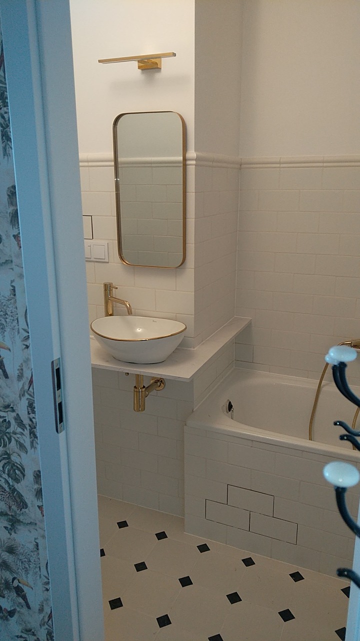 Wykończona łazienka w stylu retro z białą armaturą z elementami w kolorze złota, białe kafelki na ścianach i podłodze z czarnymi akcentami, umywalka nablatowa z baterią w kolorze złota, lustro...