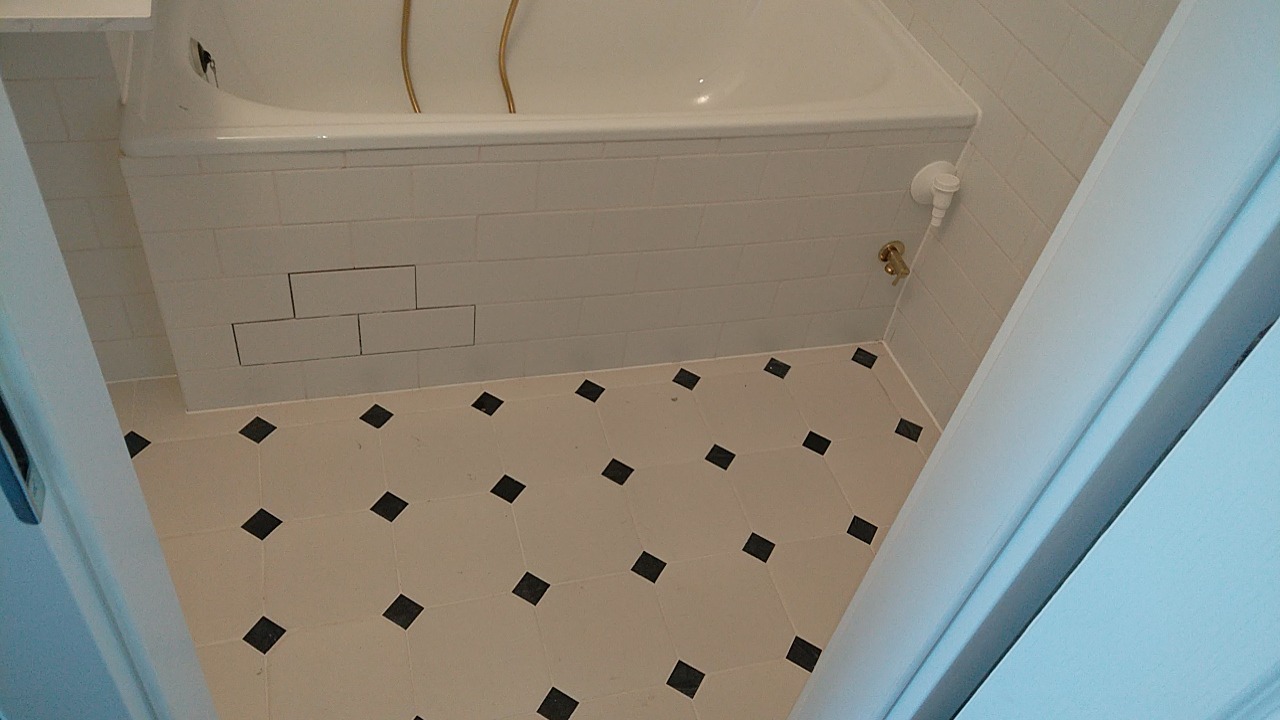 Wykończona łazienka z białą wanną obudowaną białymi, prostokątnymi płytkami, kremowa podłoga z czarnymi karo, widok zza białych drzwi.