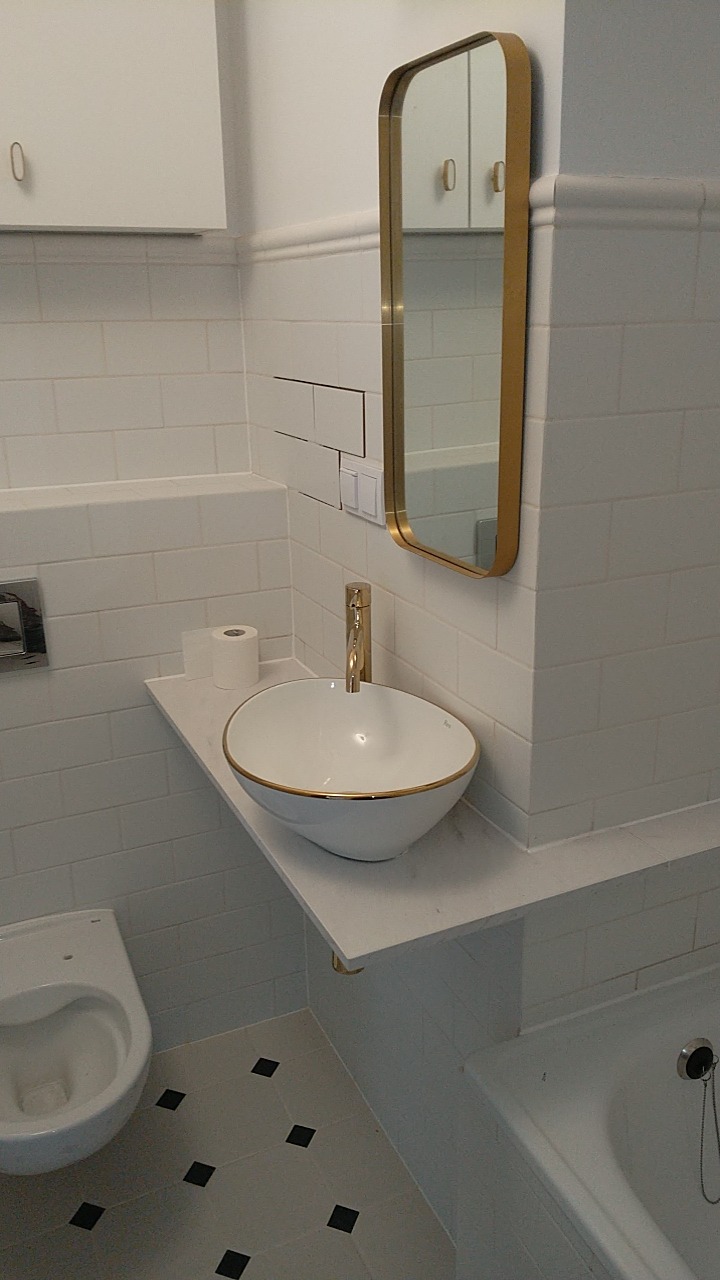 Biała łazienka z umywalką nablatową z złotym obramowaniem, baterią w kolorze złota, lustrem w złotej ramie i białymi płytkami z czarnymi akcentami na podłodze.
