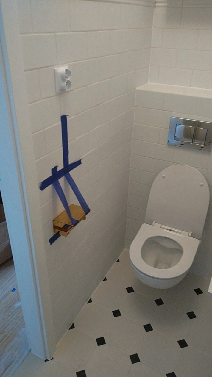 Biała łazienka z toaletą, białe płytki na ścianach i podłodze z czarnymi akcentami, gniazdko elektryczne i fragment instalacji podtynkowych zabezpieczony niebieską taśmą malarską.