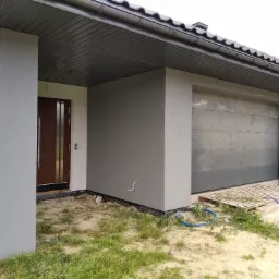 Nowoczesny dom jednorodzinny z szarą elewacją i brązowymi drzwiami wejściowymi, widok na fragment budynku z garażem, trawnik z resztkami materiałów budowlanych.