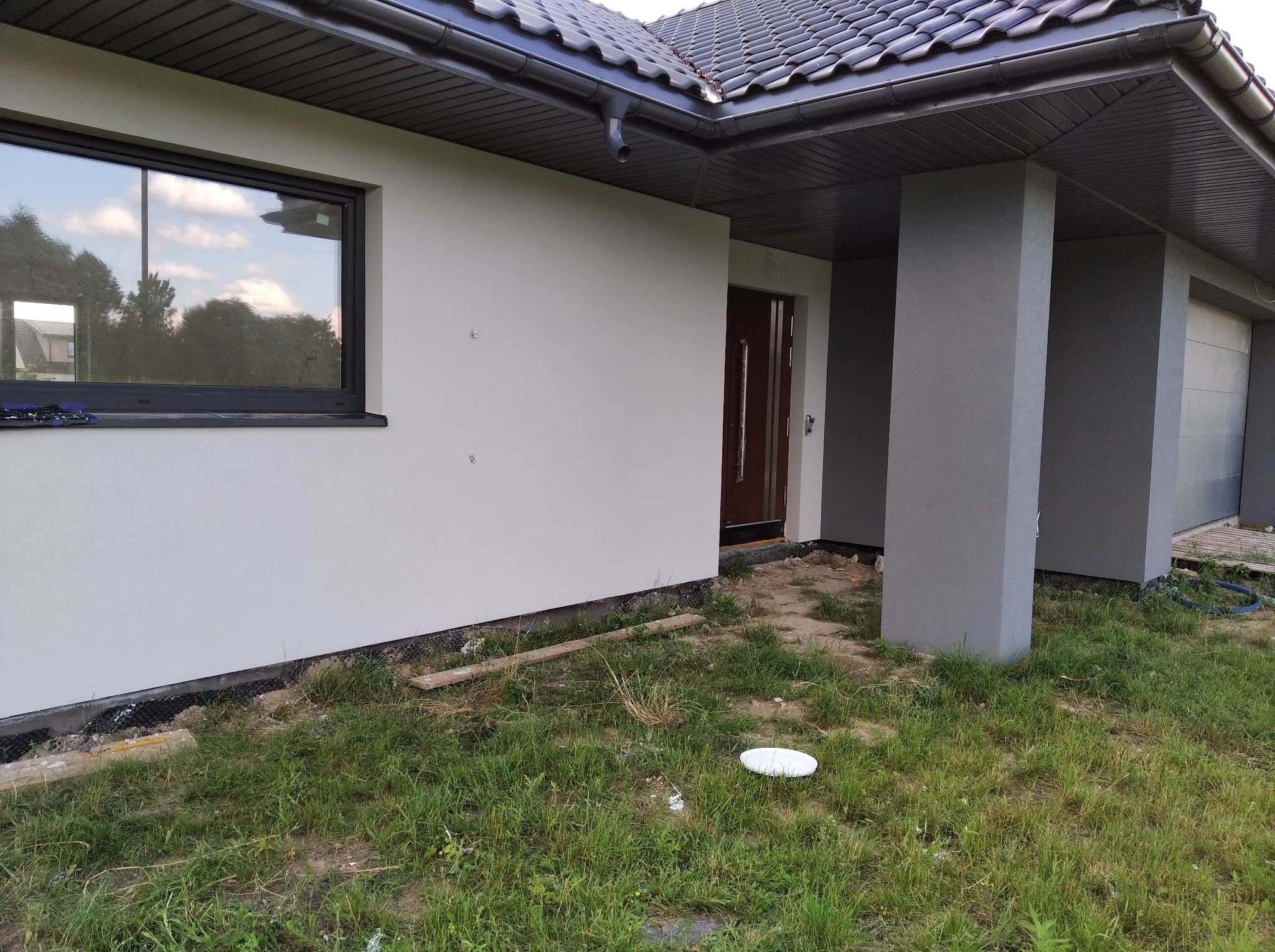 Jasna elewacja domu jednorodzinnego z ciemną stolarką okienną i grafitową dachówką, widoczne rynny i fragment trawnika z niedoskonałościami.