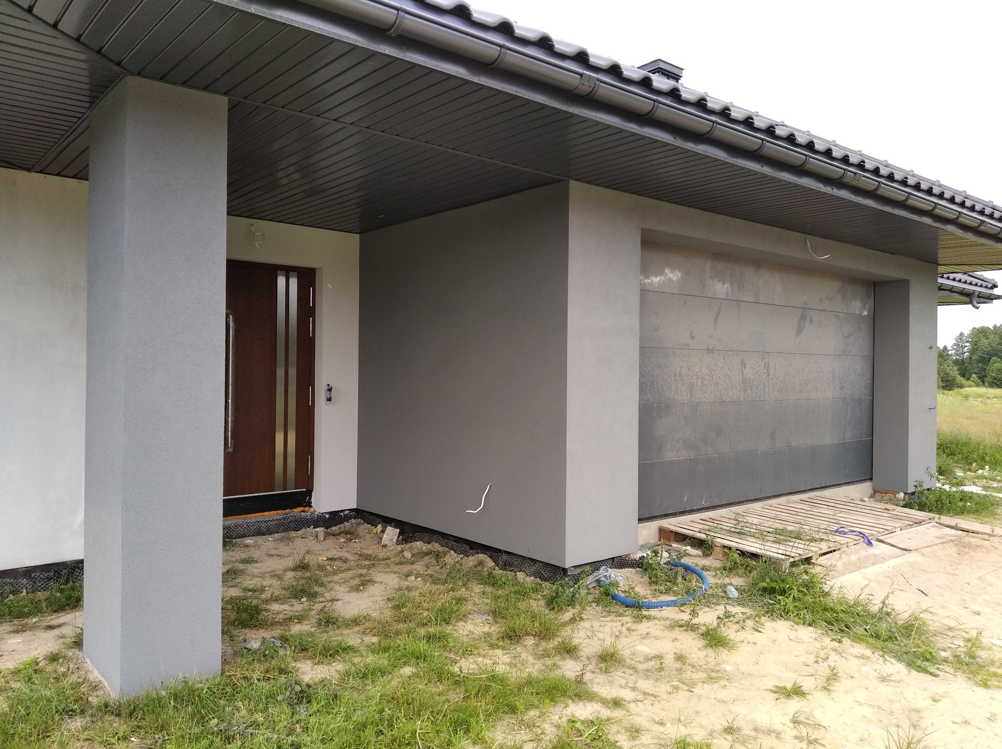 Nowoczesny dom jednorodzinny z szarą elewacją i brązowymi drzwiami wejściowymi, widok na fragment budynku z garażem, trawnik z resztkami materiałów budowlanych.