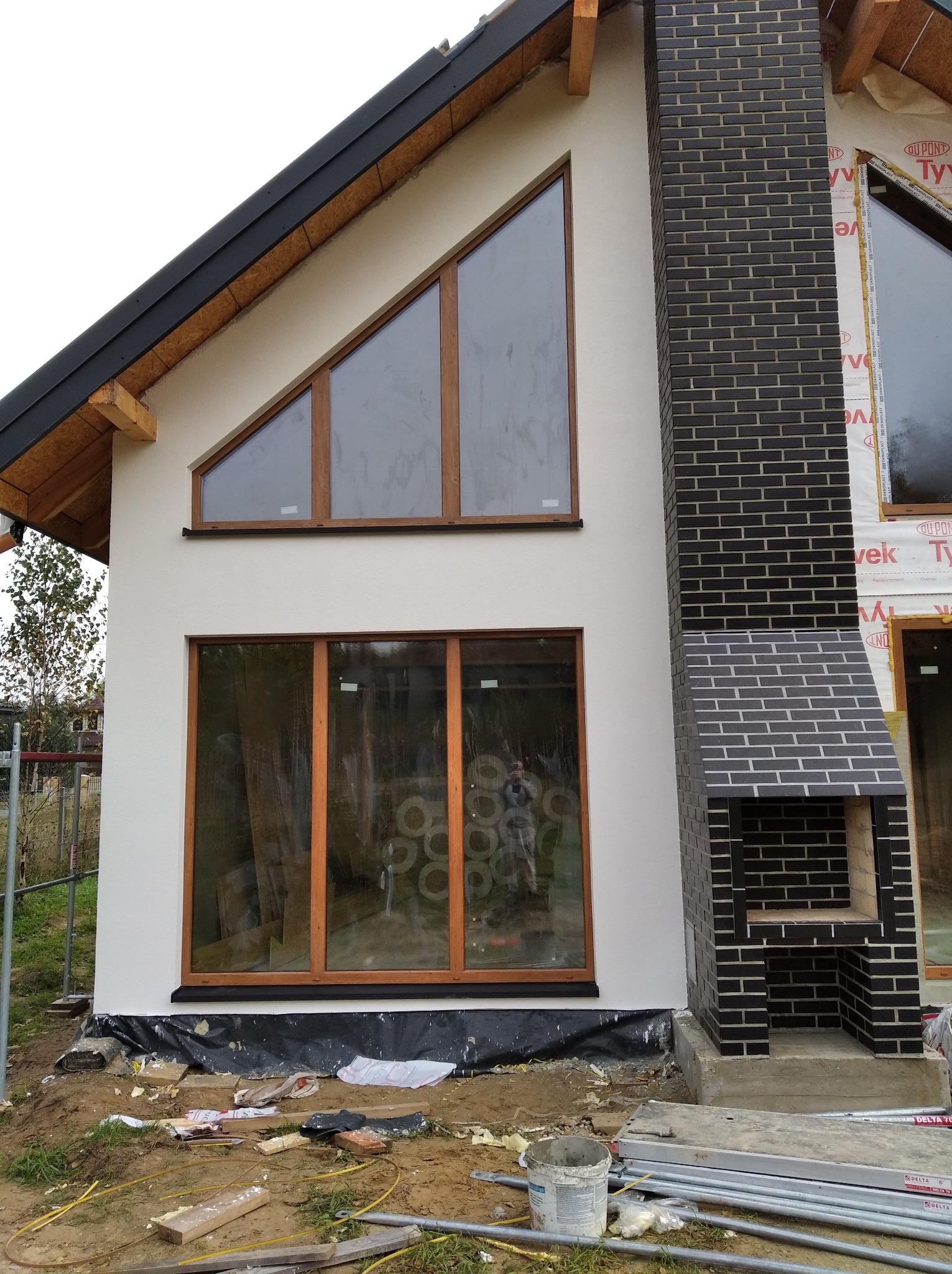 Fragment fasady domu jednorodzinnego z oknami o drewnianych ramach i kominem z ciemnej cegły, w trakcie prac wykończeniowych.
