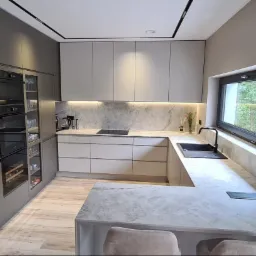 Nowoczesna kuchnia w odcieniach szarości z marmurowym blatem i oknem z widokiem na zieleń. Oświetlenie LED podkreśla elegancki design. Zabudowa z piekarnikiem i barkiem.