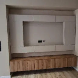 Nowoczesna zabudowa ściany TV z beżowymi szafkami i drewnianą komodą. Minimalistyczny design, gładkie fronty, widoczne gniazdka. Jasne wnętrze, parkiet.