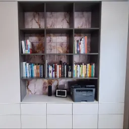 Biała zabudowa meblowa z szarymi akcentami, p&oacute;łkami na książki i marmurkowym tłem. Na blacie drukarka, głośnik i ramka. Minimalistyczny design, nowoczesne wykończenie.