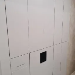 Nowoczesna łazienka: białe szafy wnękowe z ukrytymi uchwytami, czarny przycisk spłuczki i podwieszana toaleta. Minimalistyczny design, jasne wnętrze.