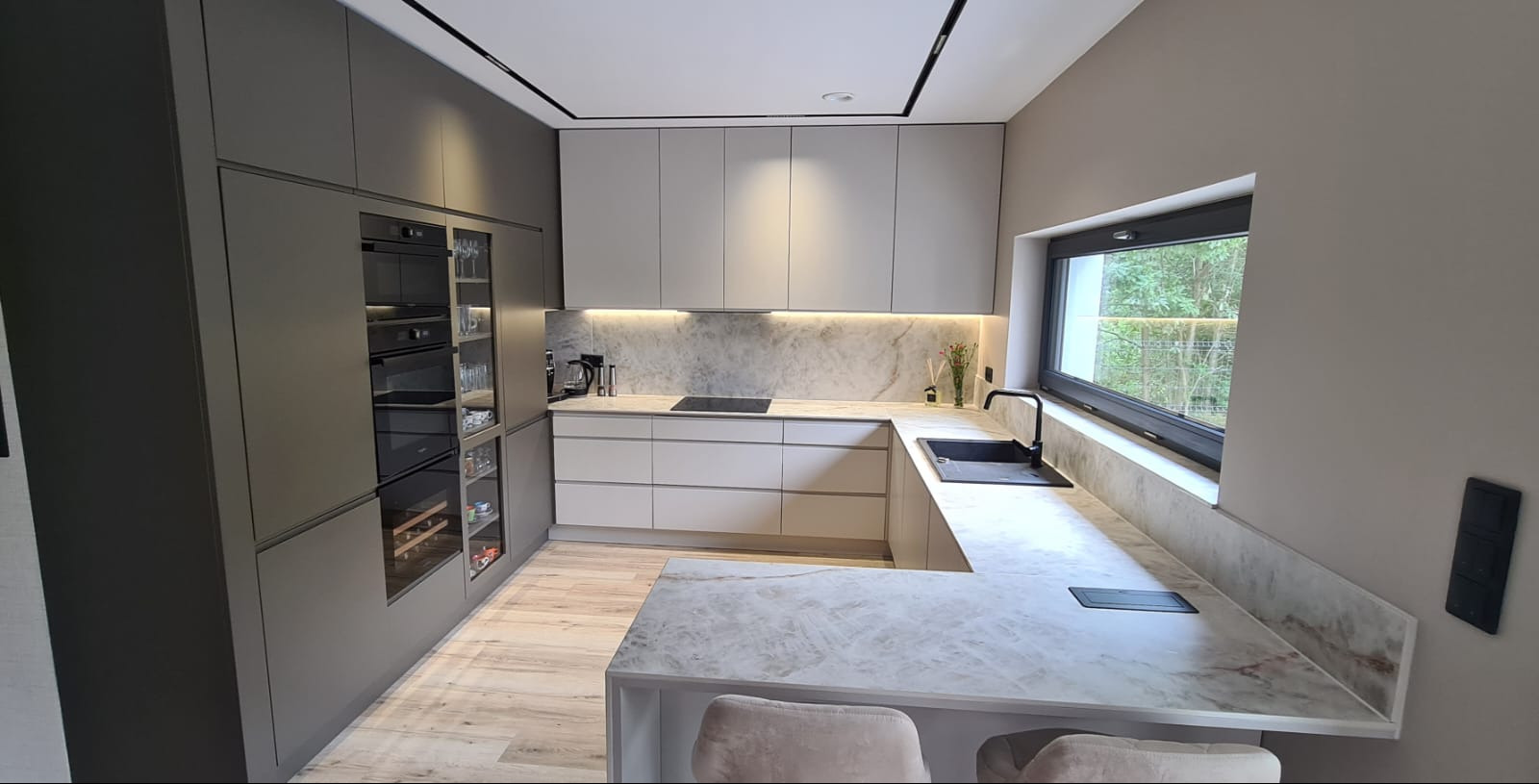 Nowoczesna kuchnia w odcieniach szarości z marmurowym blatem i oknem z widokiem na zieleń. Oświetlenie LED podkreśla elegancki design. Zabudowa z piekarnikiem i barkiem.