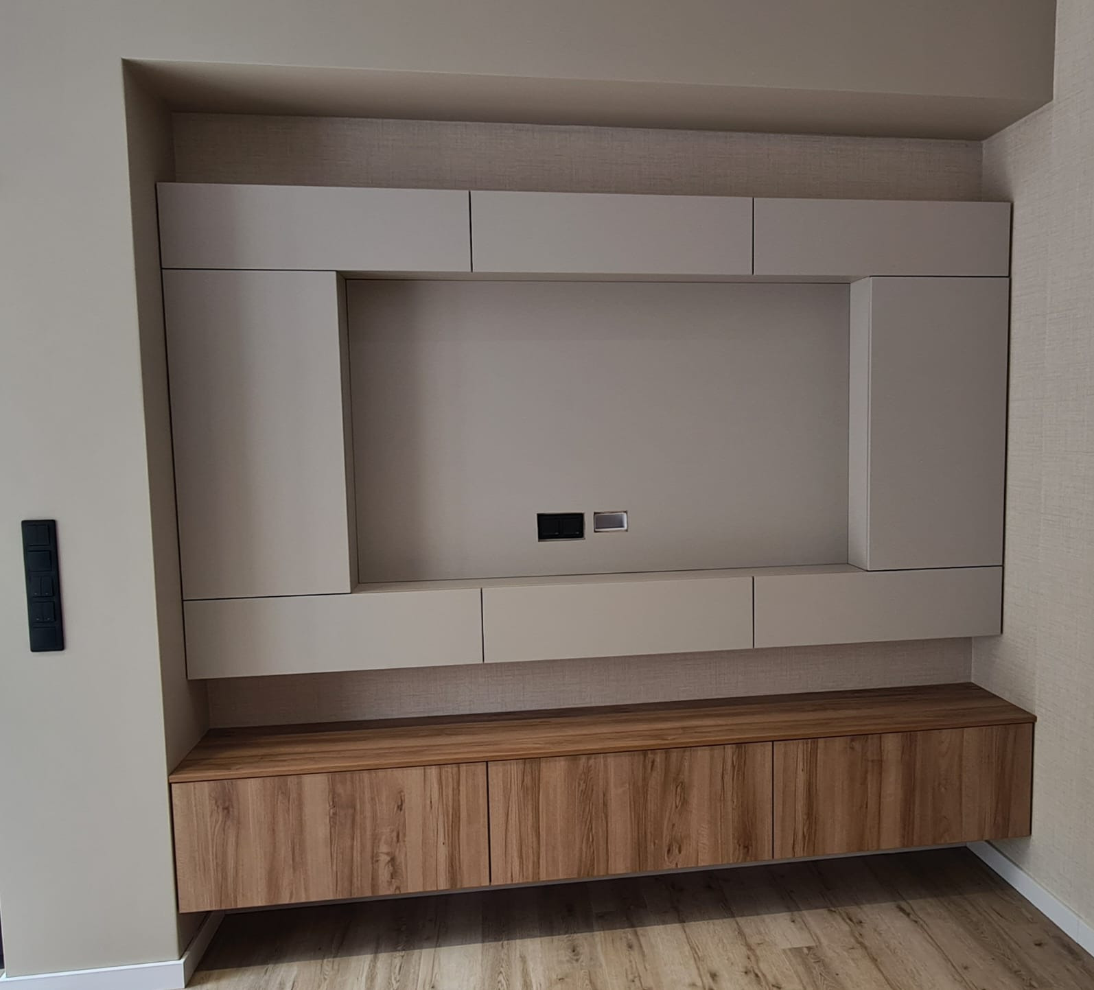 Nowoczesna zabudowa ściany TV z beżowymi szafkami i drewnianą komodą. Minimalistyczny design, gładkie fronty, widoczne gniazdka. Jasne wnętrze, parkiet.