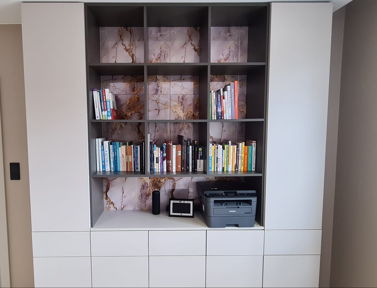 Biała zabudowa meblowa z szarymi akcentami, półkami na książki i marmurkowym tłem. Na blacie drukarka, głośnik i ramka. Minimalistyczny design, nowoczesne wykończenie.