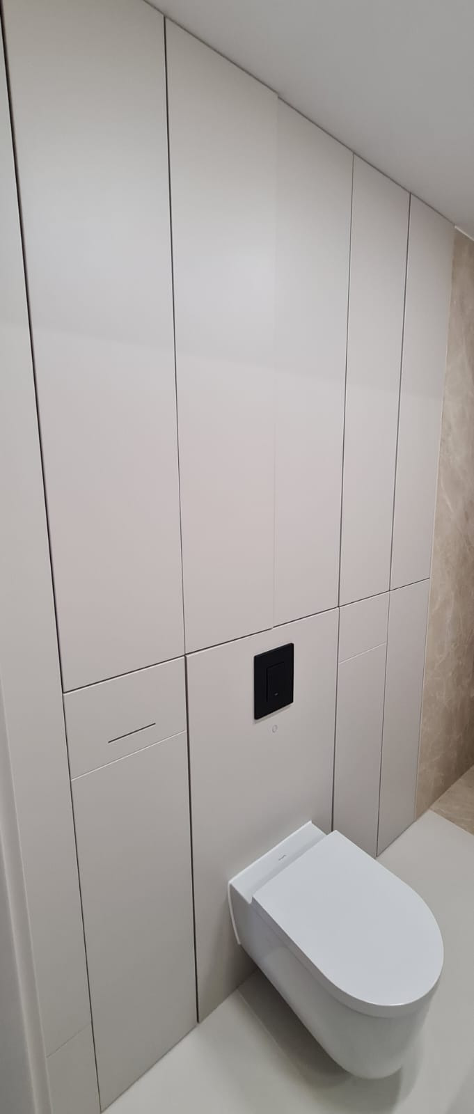 Nowoczesna łazienka: białe szafy wnękowe z ukrytymi uchwytami, czarny przycisk spłuczki i podwieszana toaleta. Minimalistyczny design, jasne wnętrze.