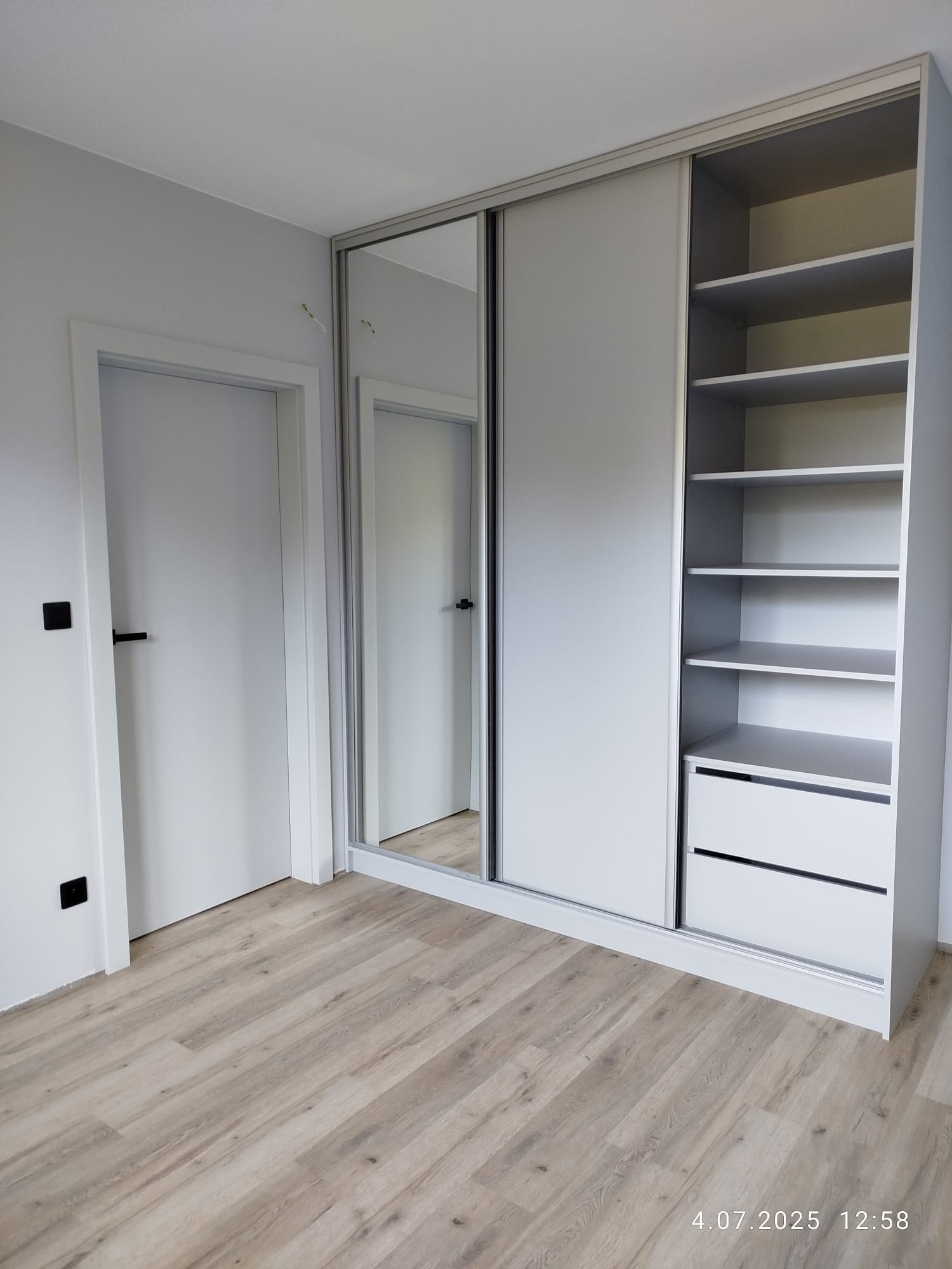 Jasnoszara szafa wnękowa z przesuwnymi drzwiami lustrzanymi i półkami, w minimalistycznym wnętrzu z drewnianą podłogą i białymi drzwiami.