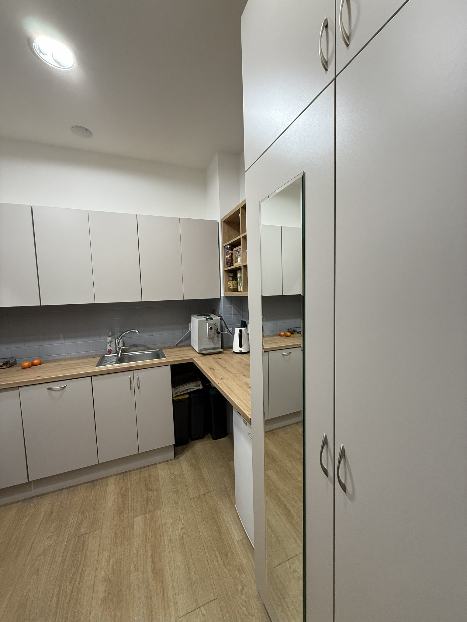 Jasna kuchnia z szarymi szafkami i drewnianym blatem, widziana z perspektywy pod kątem. Na blacie ekspres do kawy i owoce. W tle płytki i regał z zapasami.